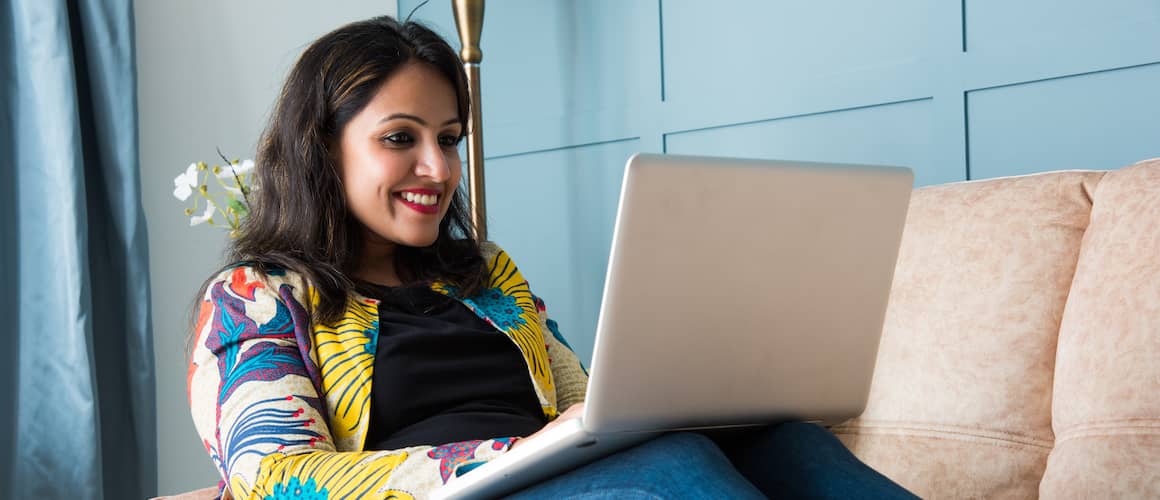 Indian woman smiling while using her laptop on the couch.