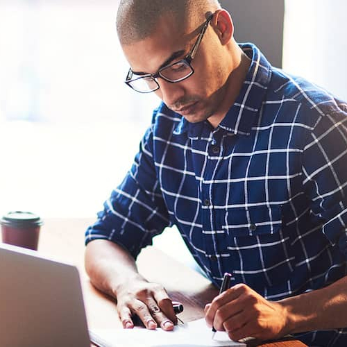 Focused man at desk writing down notes while desktop and laptop.