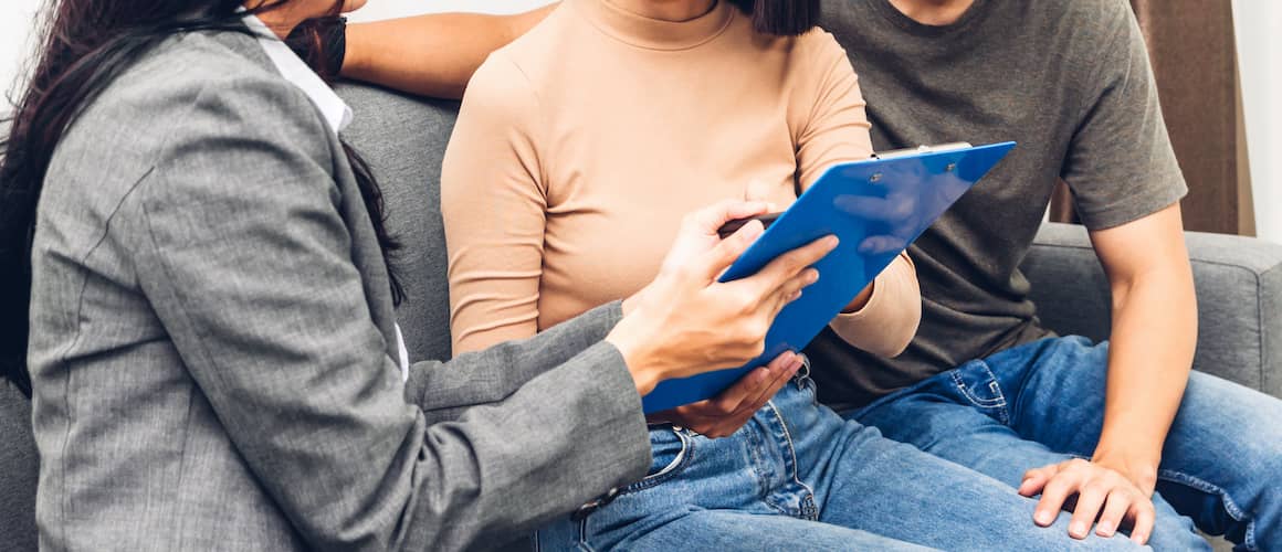 Real estate agent presenting paper documents to a couple on a couch, potentially discussing property details.