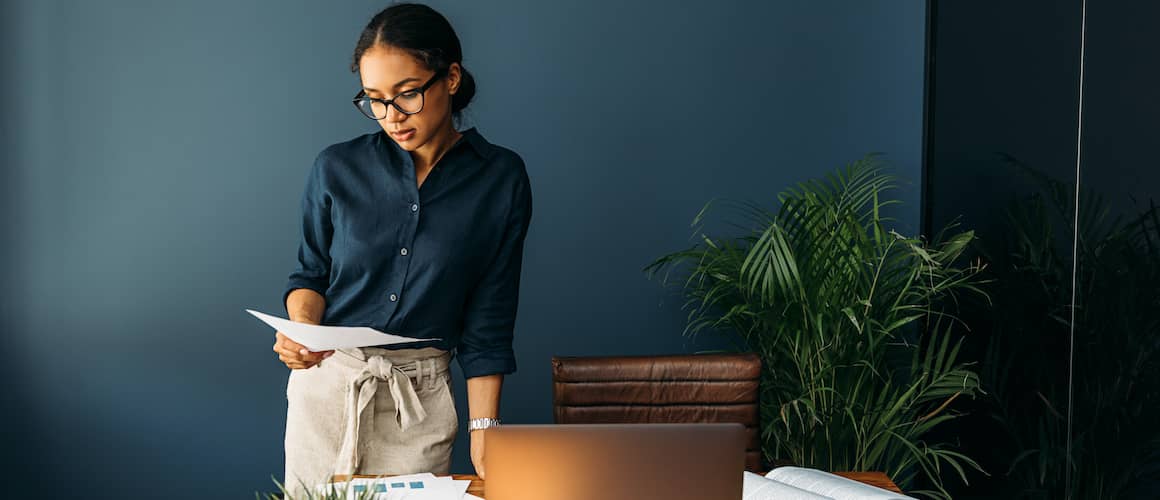 A woman wearing glasses stands at a desk, working on a laptop computer and looking on a piece of paper.