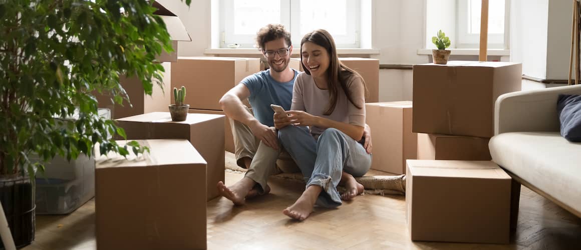 A young couple sits on the floor next to cardboard boxes, possibly discussing or planning their move into a new home.