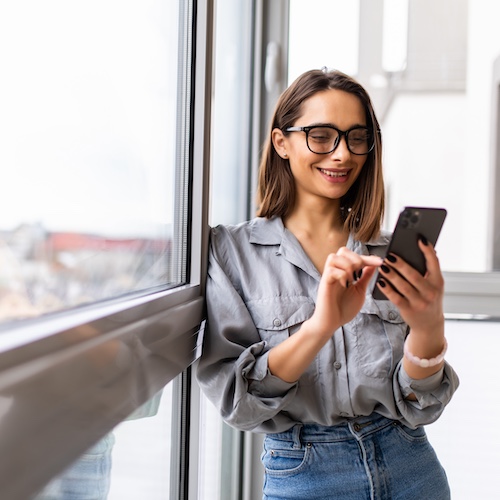 Young woman wearing glasses and using her cell phone while leaning against a large window.