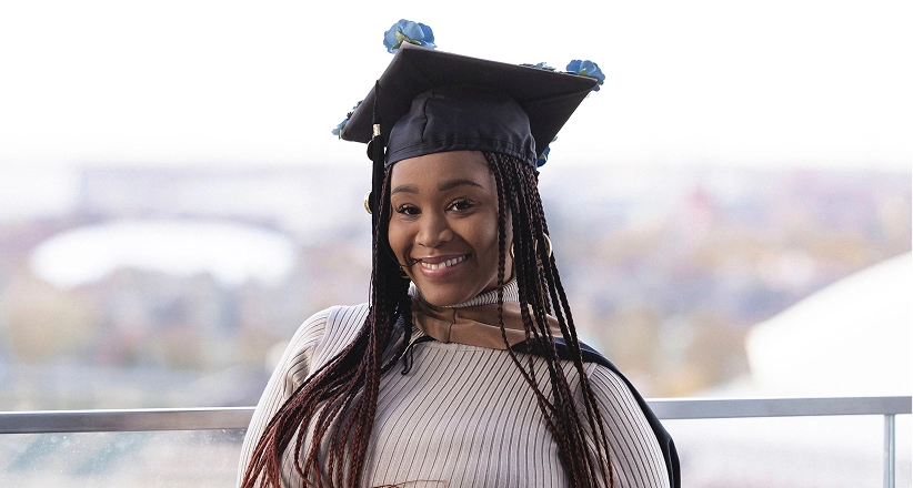 A women with long hair is wearing a graduation cap and standing in front of a window with an outdoor view.