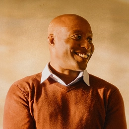 A smiling bald man in a brown sweater layered over a white collared shirt looks off to the side against a warm, neutral background, appearing relaxed and cheerful.