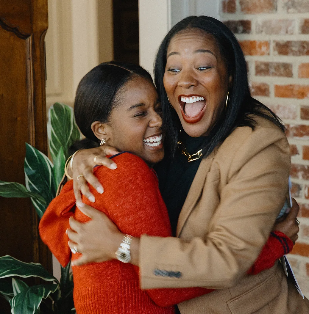 A Black mom in a tan blazer hugs her teenage daughter, sharing excitement over the new home they just bought with a jumbo loan from Rocket Mortgage.