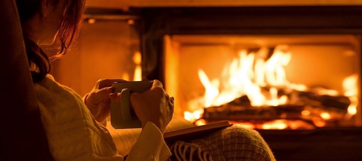 RHB Assets From IGX: Woman reading a book by a cozy fireplace, wrapped in a warm blanket.