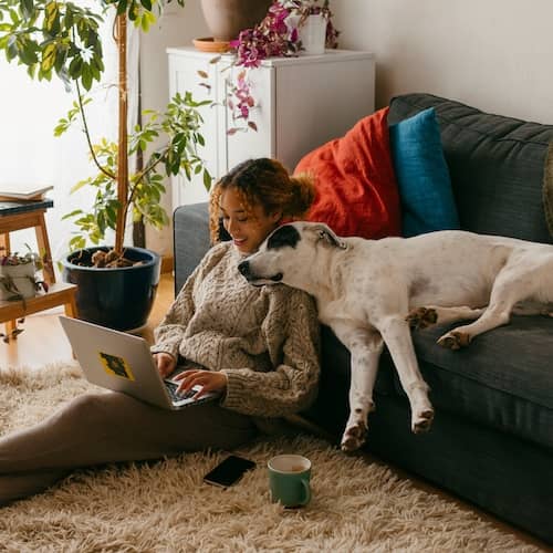 A young woman working on a laptop from the floor of her living room while her dog sleeps on the couch with its head on her shoulder.