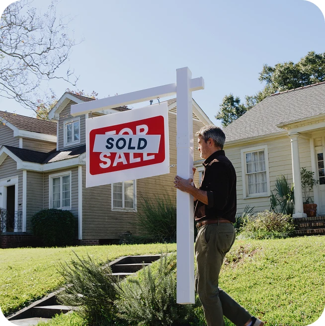 A man stands in the backyard of a modest home, carefully pulling a 'Sold' sign out of the grass.
