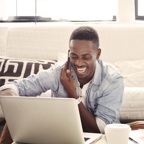 Man smiling while speaking on phone, looking at laptop in living room.