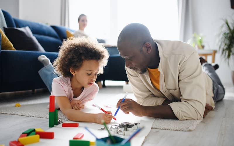 Father and daughter drawing on paper with markers on the floor of the living room with the mother on the couch in the background smiling.