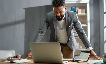 A smiling black man wearing a business jacket is standing at a desk, looking down at a laptop and with a calendar diary sitting open in front of him.