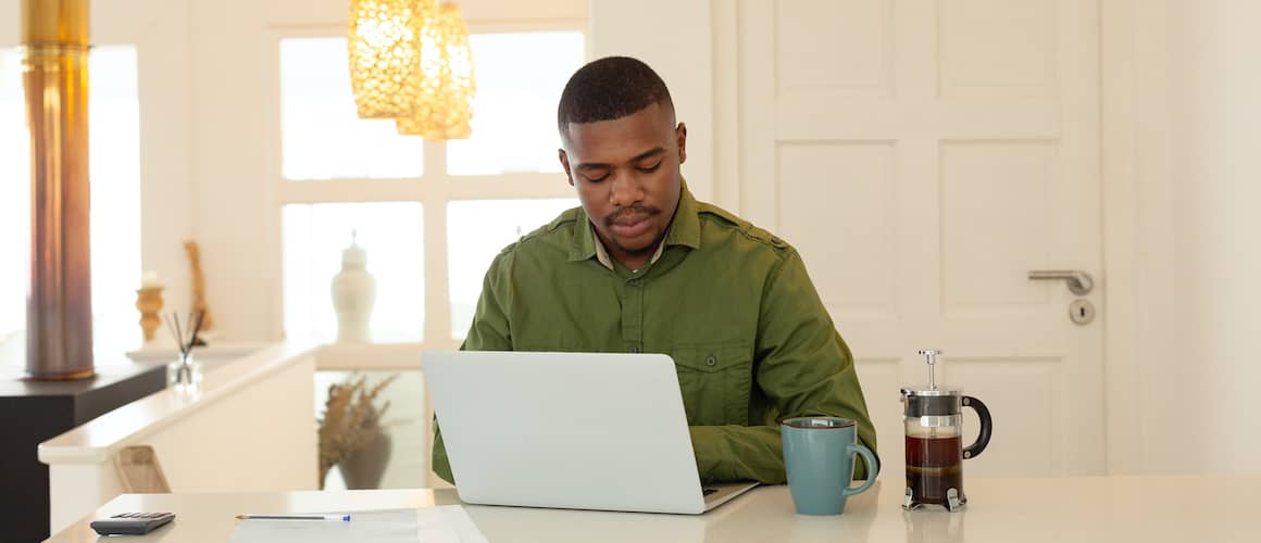 A man in green sitting at a counter with a French press and a laptop.