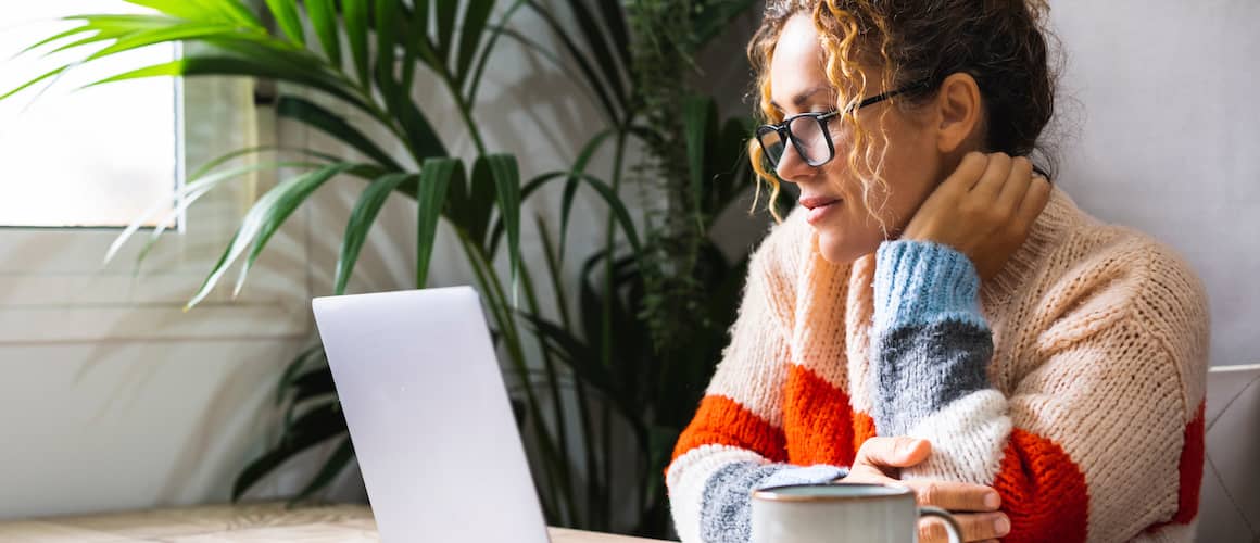 Young adult woman with curly hair and dark glasses, sitting at table with mug of coffee, looking at computer.