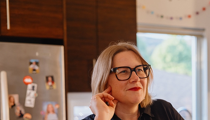 Woman with red lipstick and black glasses standing in kitchen smiling.
