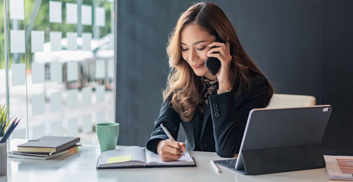 Young Asian woman talking on the phone, writing notes down in her notebook.