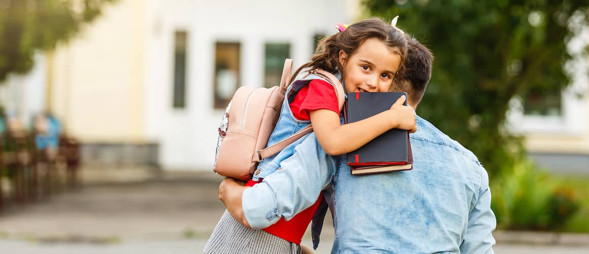 Father carrying daughter with backpack on into school.