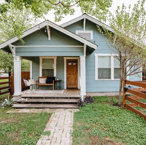Small blue bungalow with porch surrounded by rustic wooden fence.
