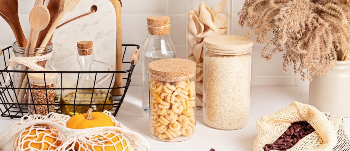 Kitchen countertop with wooden utensils, glass canisters filled with dry goods, reusable grocery bag with produce and dried natural element in stoneware vase.