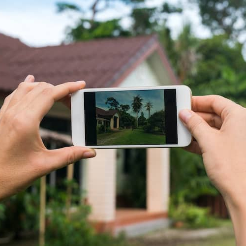 Hands holding a smartphone photographing a house, possibly related to real estate or property viewing.