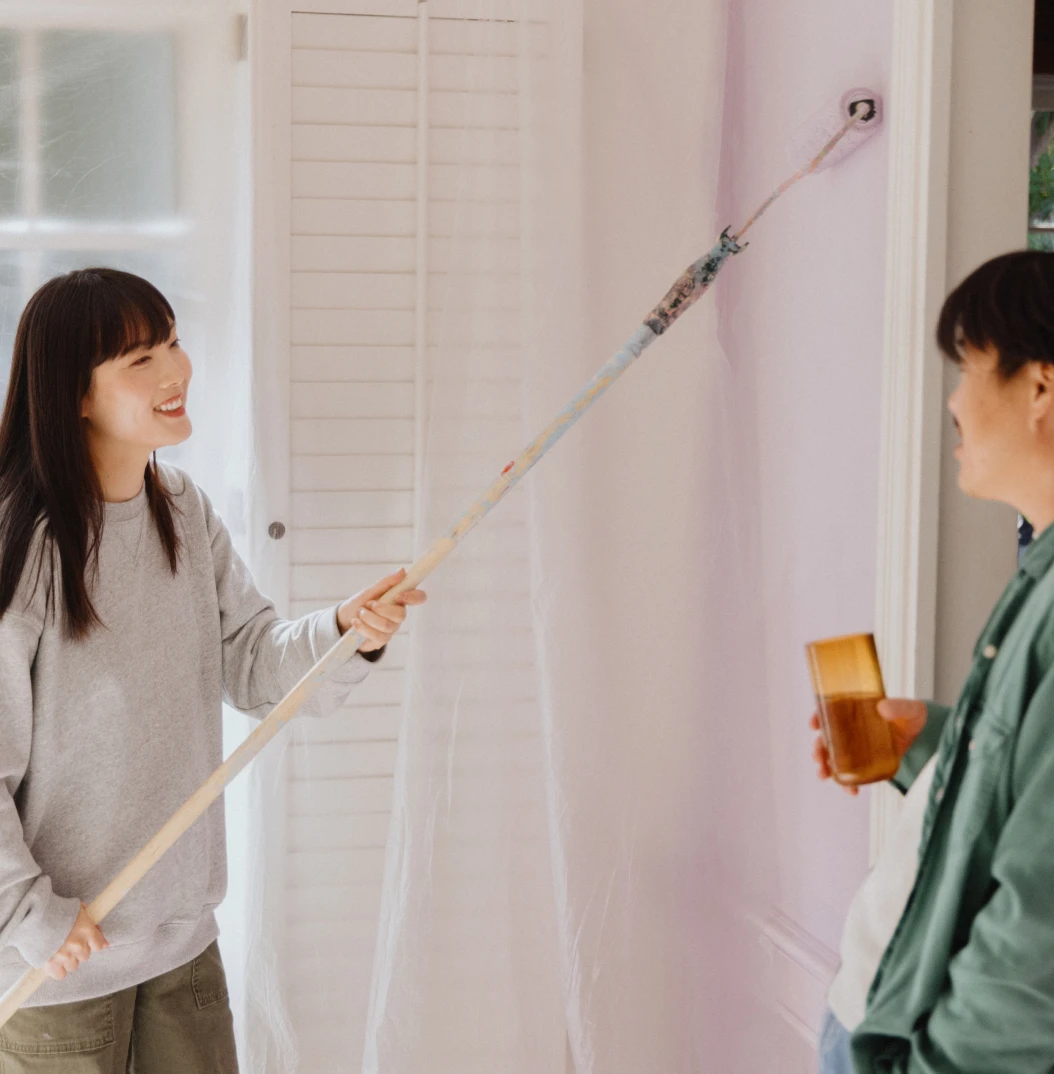 A couple paint an interior wall together, smiling as one uses a paint roller and the other looks on.