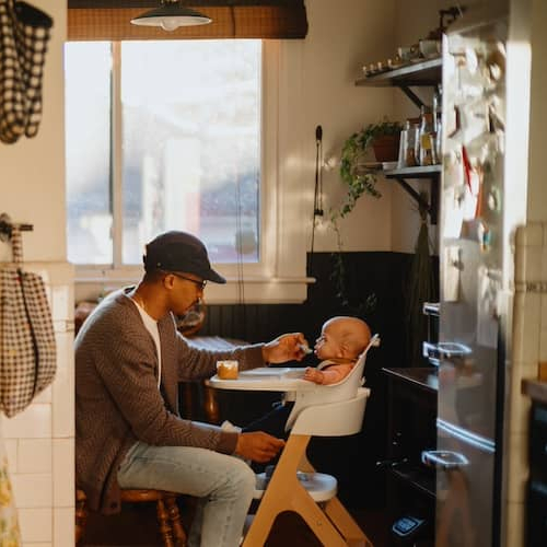 Mother in kitchen cooking while father feeds child in high chair in dining room.