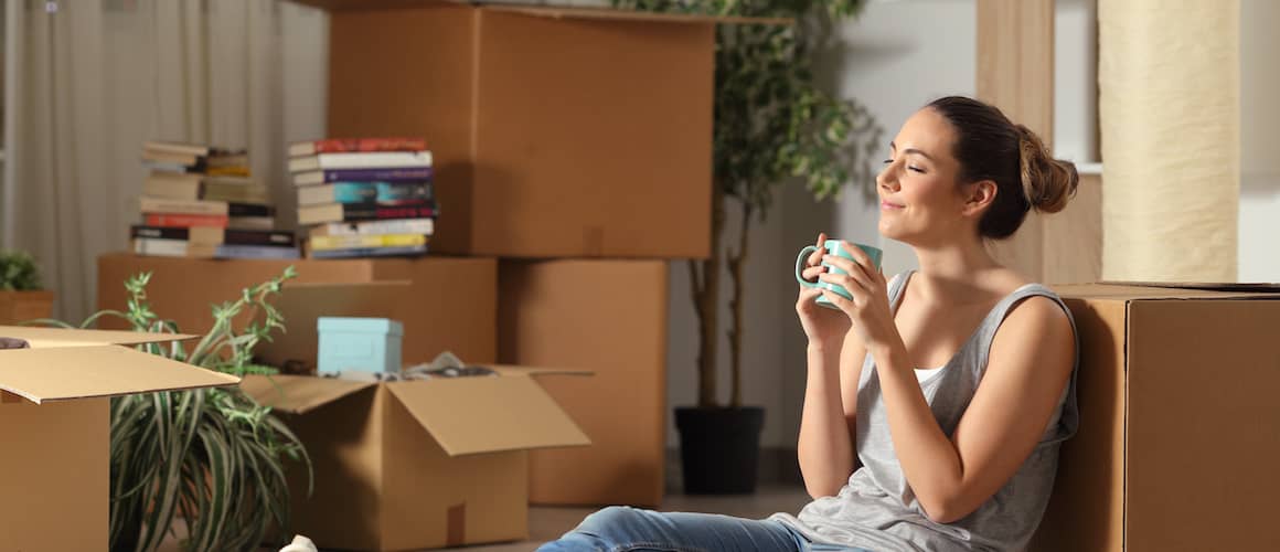 A woman smiling  alone with a cup of tea in a new house, while engaged in unpacking and shifting.