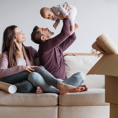 Young couple holding curious baby sitting on the couch while unpacking cardboard boxes.