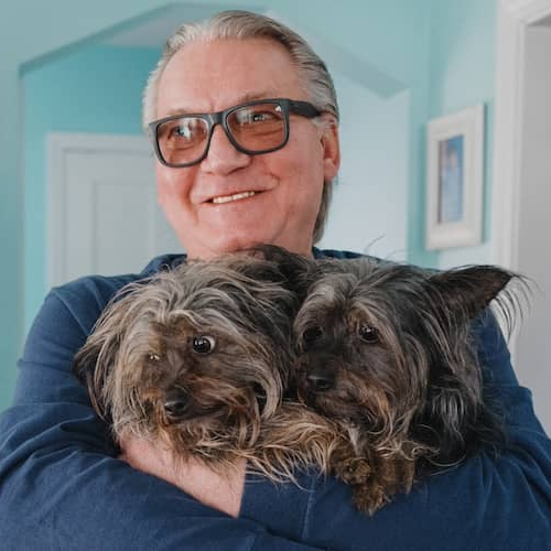 Man smiling and holding two fluffy small dogs in living room.