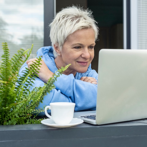 Woman with pixie cut silver hair having coffee while checking email on laptop on outdoor balcony.