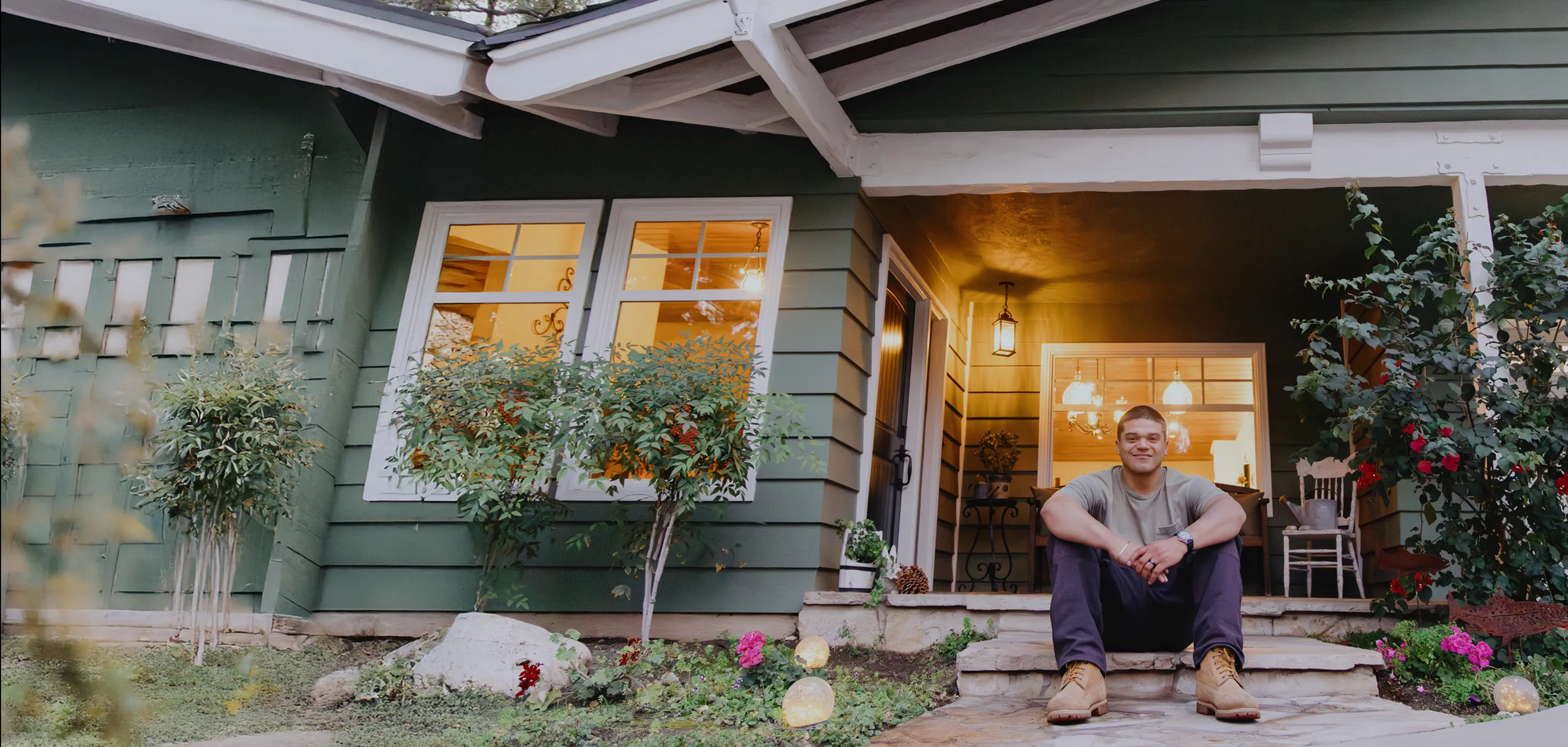 A Hispanic man wearing work boots, brown pants and a tan t-shirt smiles proudly while sitting on the front steps of a stone porch, with a lighted picture window in the background.