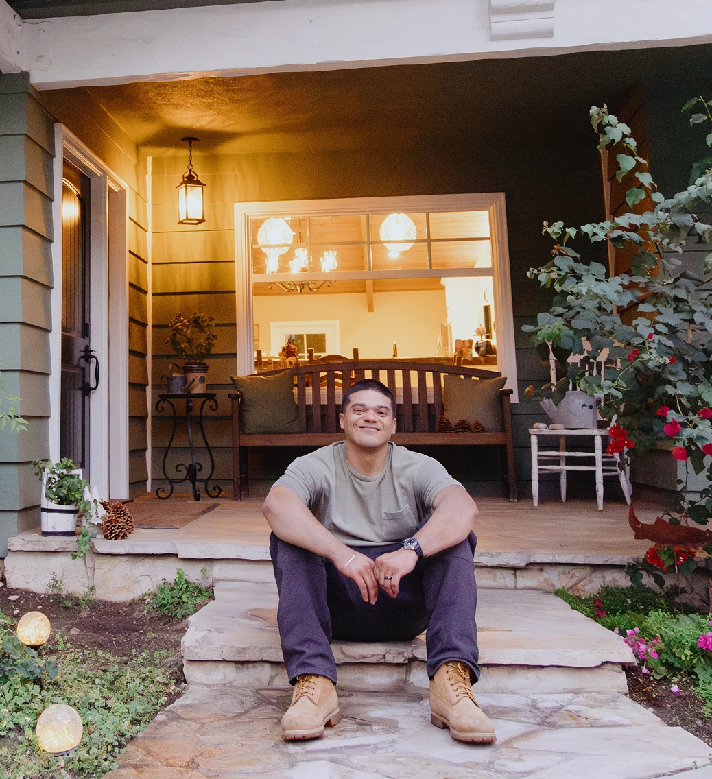 A man sits proudly, smiling on the front stoop of his green house.