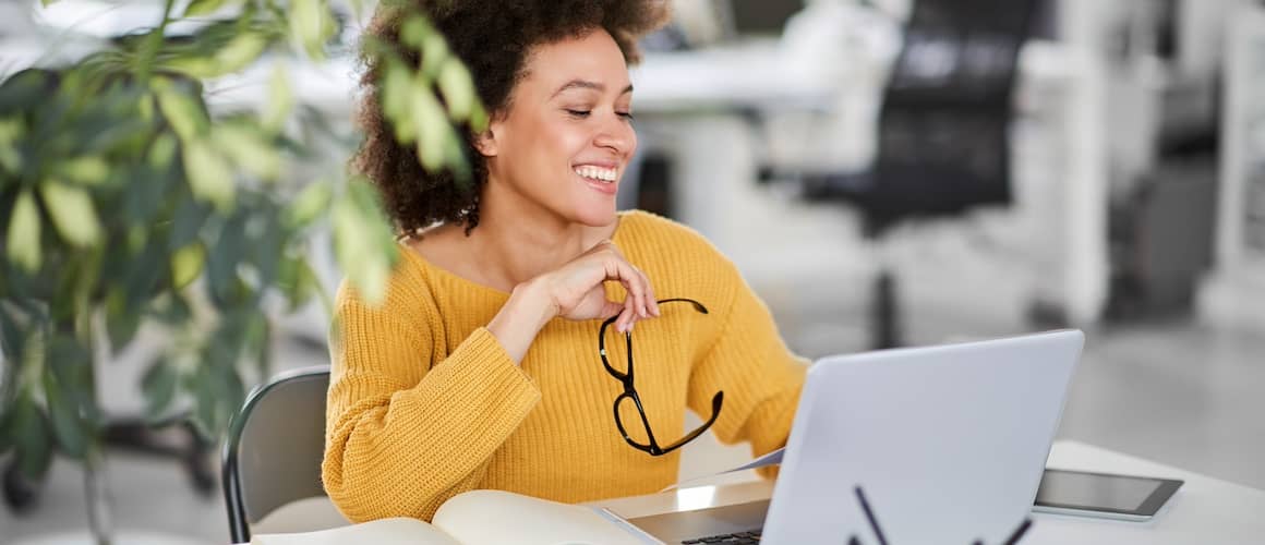 A young woman wearing yellow and holding glasses in her hand smiling while using her laptop.