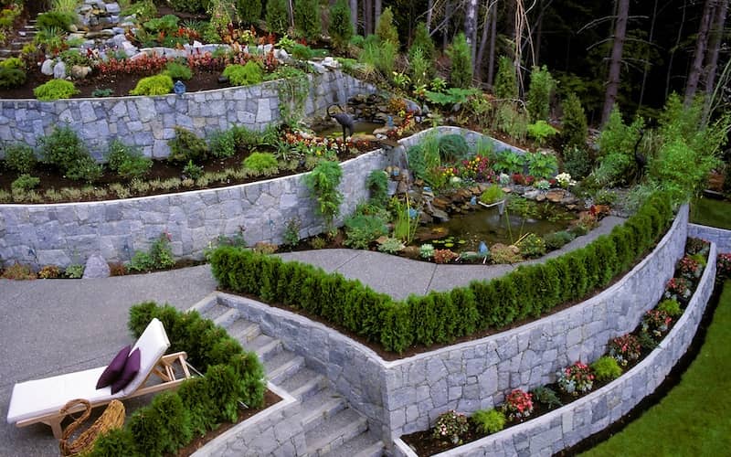 Curved stone retaining wall lined with flowers, walkways and a lounge chair. 