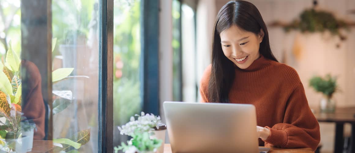 A smiling Asian woman in a clay-colored sweater working on a laptop near windows, potentially in a work or home office setting.