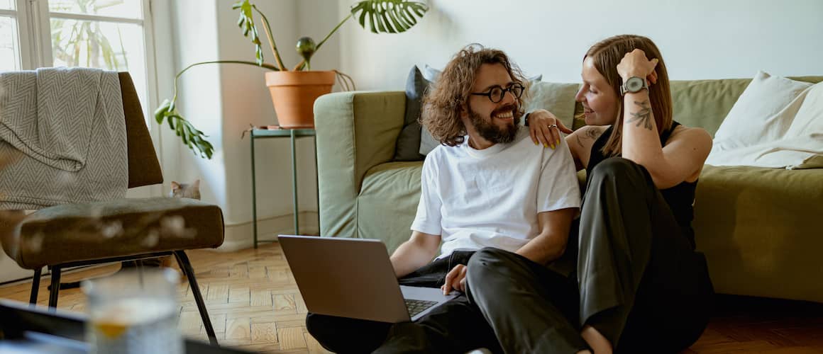 Young couple smiling while on floor of apartment looking at catalog homes on laptop.