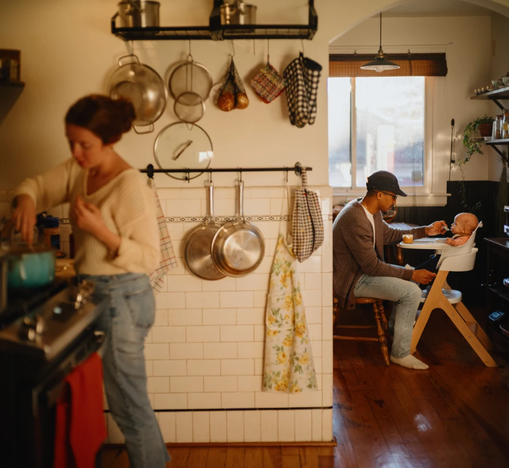 A woman is cooking at the stove in her kitchen while her husband feeds their baby in the background.