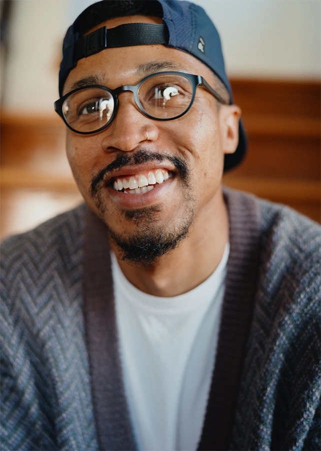 A close up portrait of a smiling black man with glasses and a hat on.