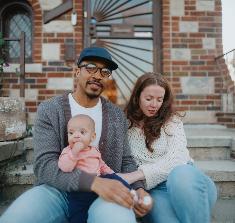 A young couple sits on the front step of their home with their baby.