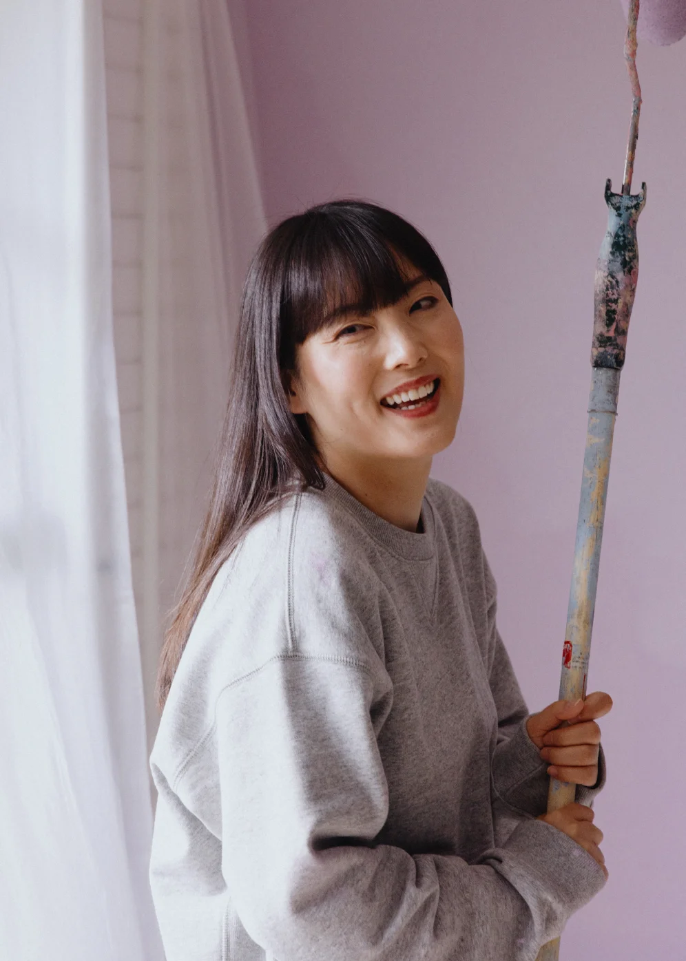 An Asian woman with long hair and bangs stands smiling in a room she is painting.