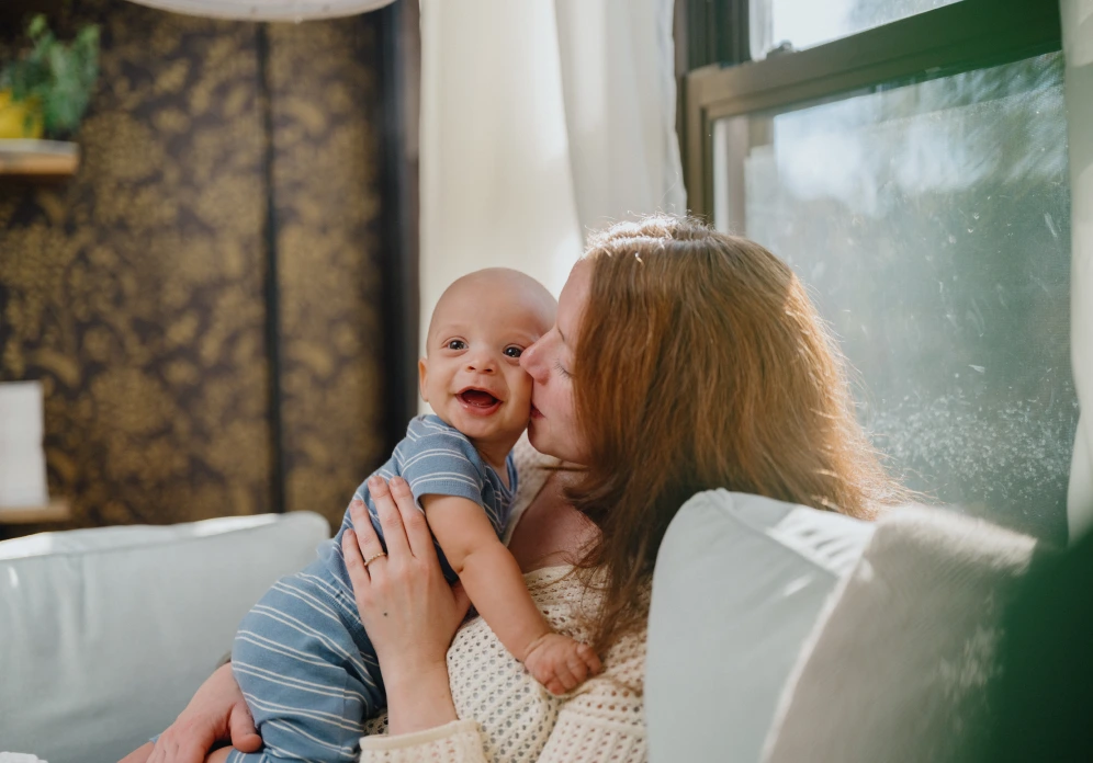 A white woman with red hair giving her baby a kiss of the cheek in their home.