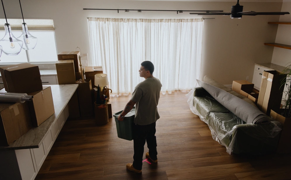 A man stands inside of his new home, holding a bin and surrounded by moving boxes.