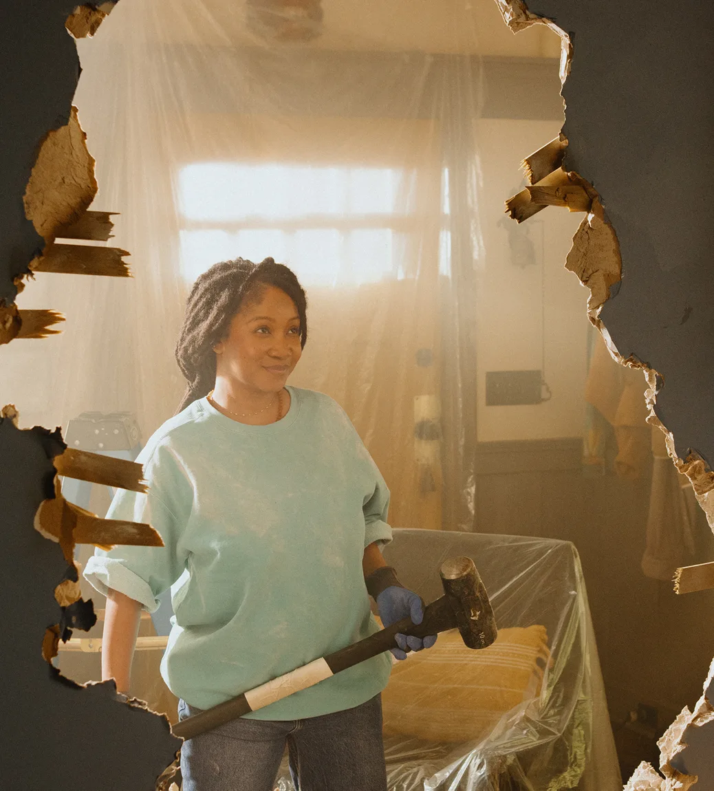 A woman in a blue sweatshirt holds a sledgehammer as she stands in front of a large hole in a wall during a home renovation.