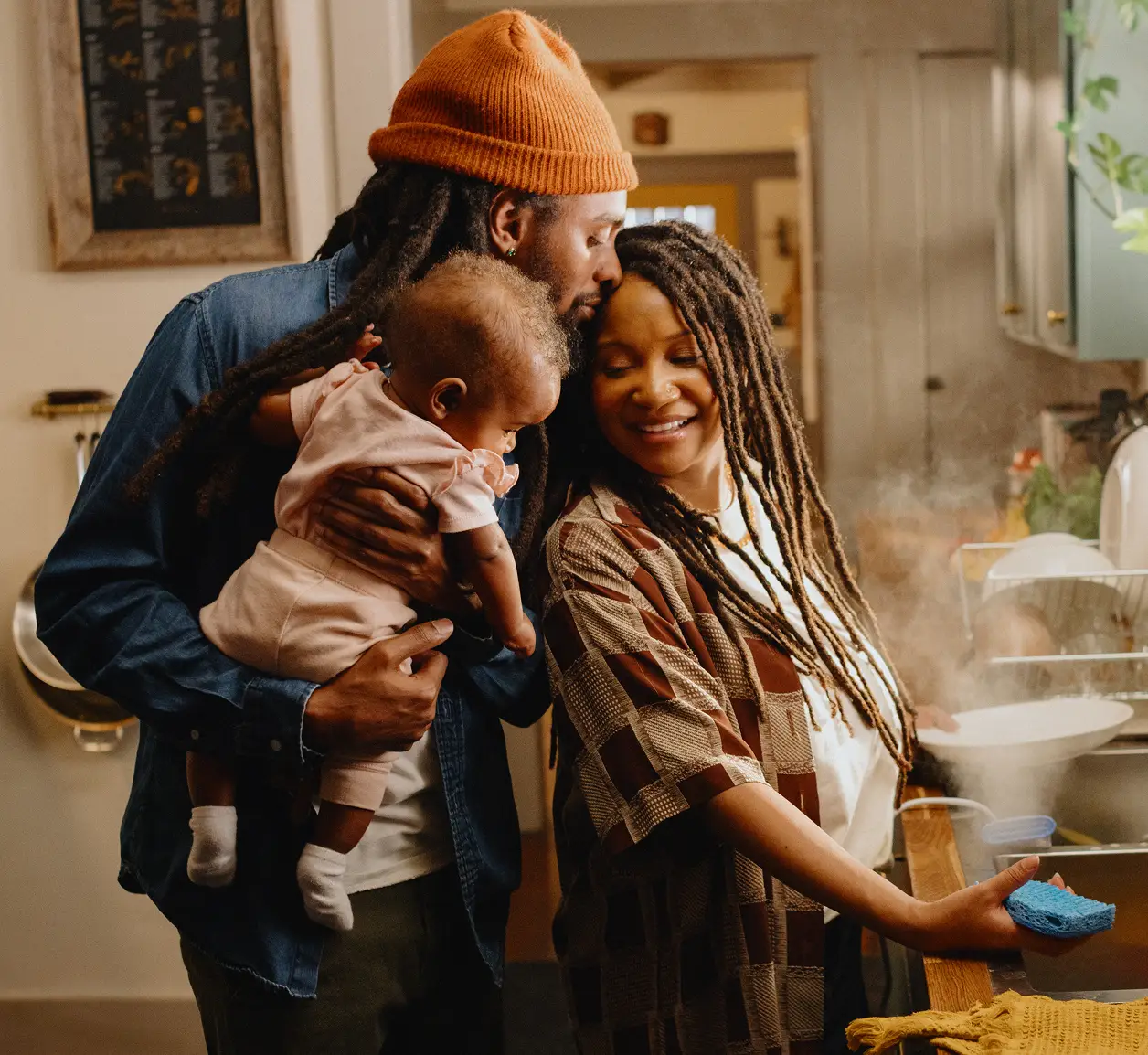 A man and woman embrace in their kitchen, while the man holds their child and the woman is cleaning dishes in the sink.