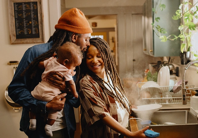 A man and woman embrace in their kitchen, while the man holds their child and the woman is cleaning dishes in the sink.