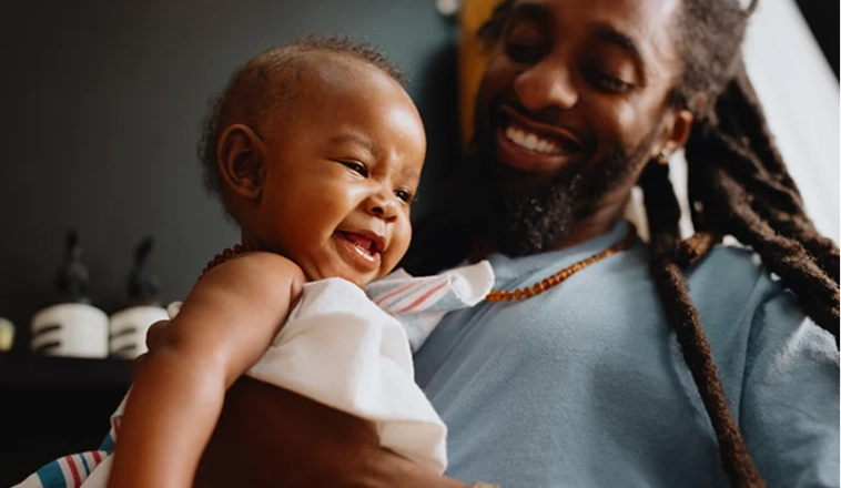 A man smiles while holding his happy baby wrapped in a blanket.