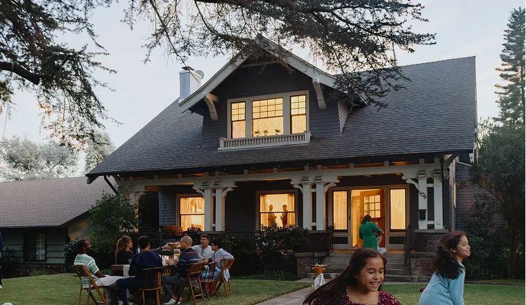 Exterior of a home in the evening, with lights on inside and a family gathering outside.