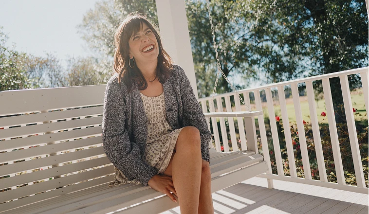 A woman smilles while sitting on the porch swing in front of her home