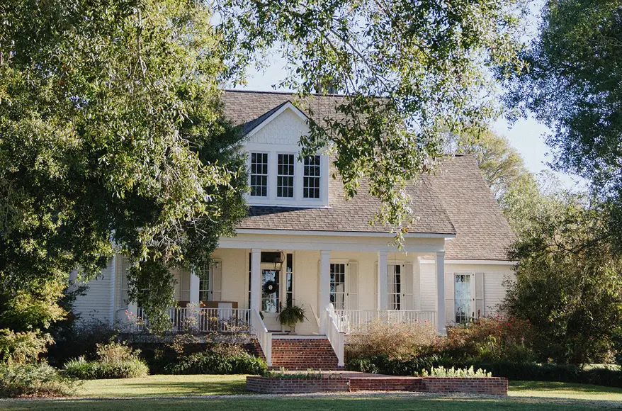 Leafy branches hang in front of a white house, with a brick walkway leading to a large covered porch.