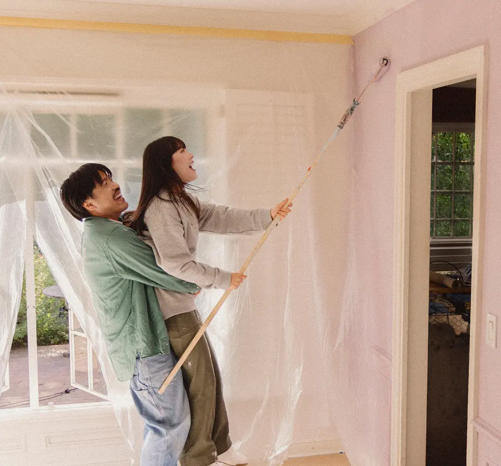 A young couple laughs together as the man lifts up the woman, who is painting the wall with a long roller.