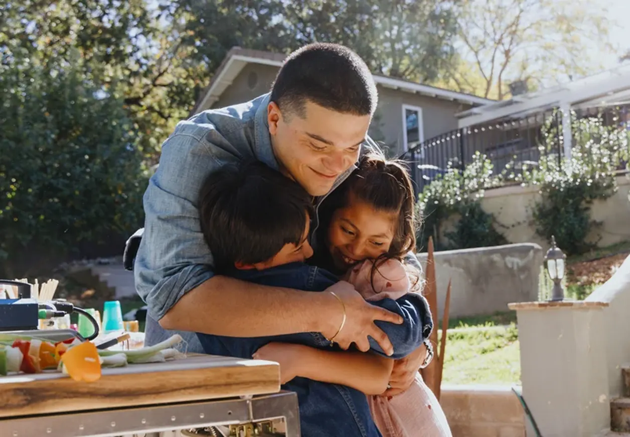 A man with short dark hair smiles and leans over to hug two kids while standing outside in the yard.